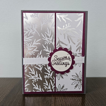 A handmade greeting card with a silver and white botanical pattern, featuring a central scalloped burgundy circle with the words 'Season's Greetings' and a white ribbon across the middle, displayed on a wooden surface against a light gray background.