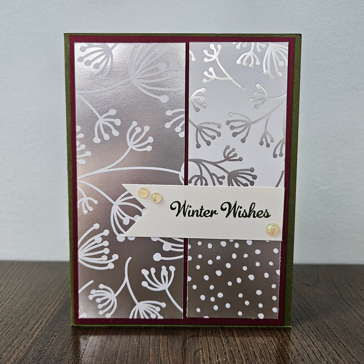 A handmade greeting card with a winter-themed design, featuring a silver and white floral pattern, a white banner with 'Winter Wishes' in black script, and small gold sequins, standing on a wooden surface against a light gray wall.