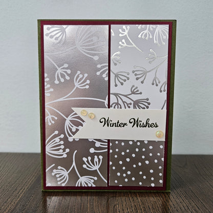 A handmade greeting card with a winter-themed design, featuring a silver and white floral pattern, a white banner with 'Winter Wishes' in black script, and small gold sequins, standing on a wooden surface against a light gray wall.