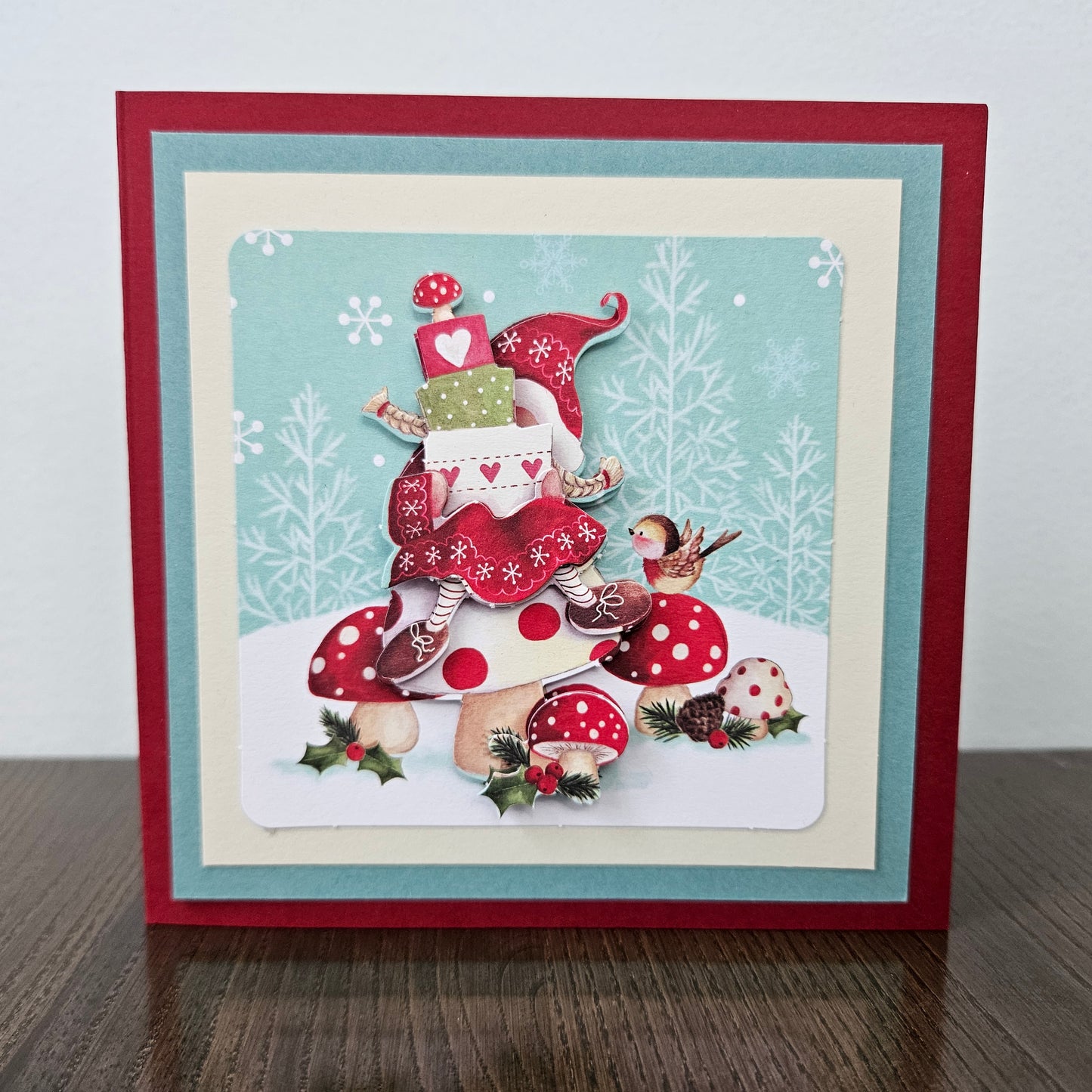 A square handmade Christmas greeting card displayed on a dark wooden surface against a light gray wall. The card features a festive illustration of a small elf character wearing a red hat and dress, surrounded by red and white polka-dotted mushrooms, pinecones, holly, and a bird, set against a light blue winter-themed background.