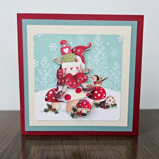 A square handmade Christmas greeting card displayed on a dark wooden surface against a light gray wall. The card features a festive illustration of a small elf character wearing a red hat and dress, surrounded by red and white polka-dotted mushrooms, pinecones, holly, and a bird, set against a light blue winter-themed background.