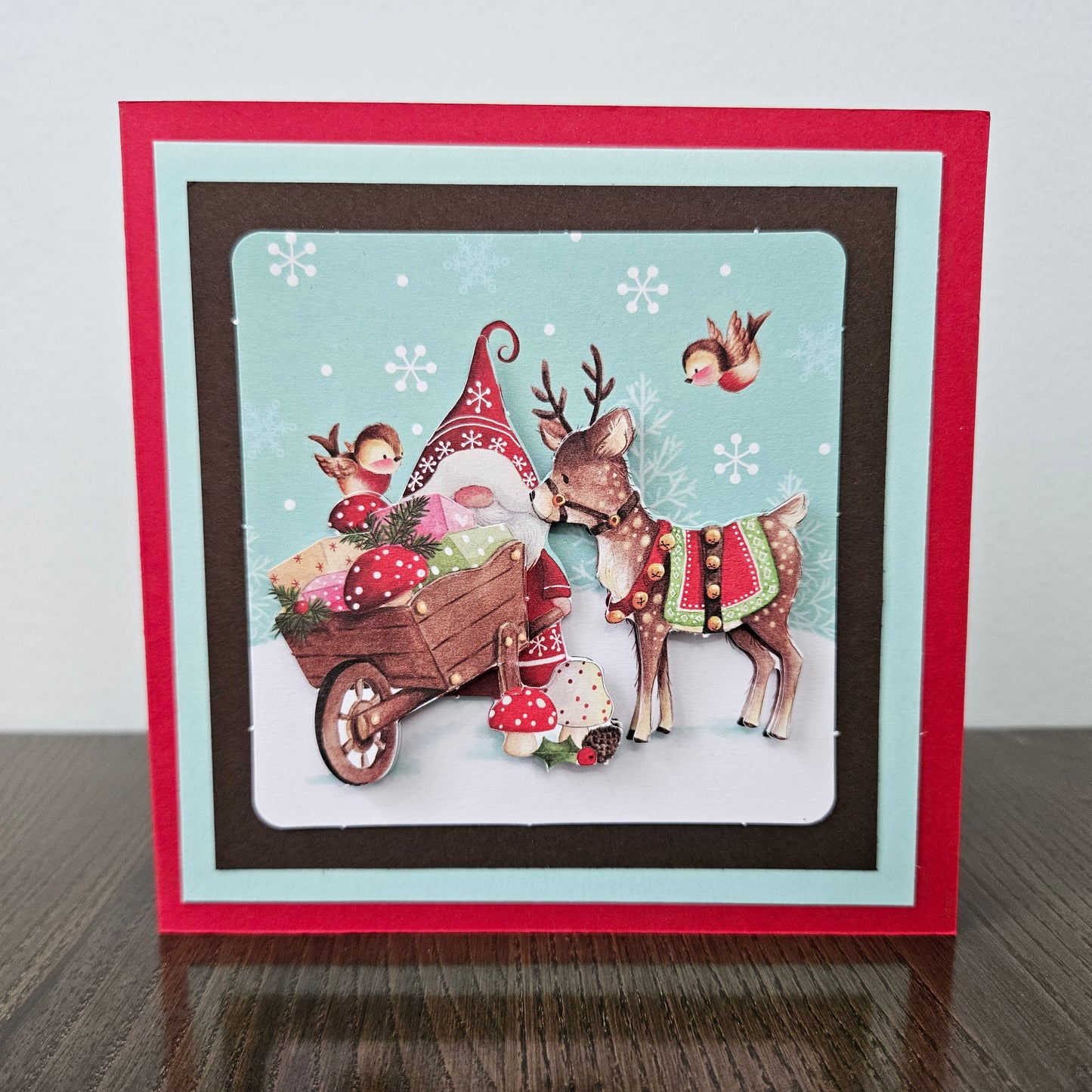 A square handmade Christmas card displayed on a dark wooden surface against a light gray wall. The card features a layered border in red, light blue, and dark brown, with a central illustration of a festive scene including a gnome in a red hat, a decorated deer, a bird, and a small cart with mushrooms and berries.