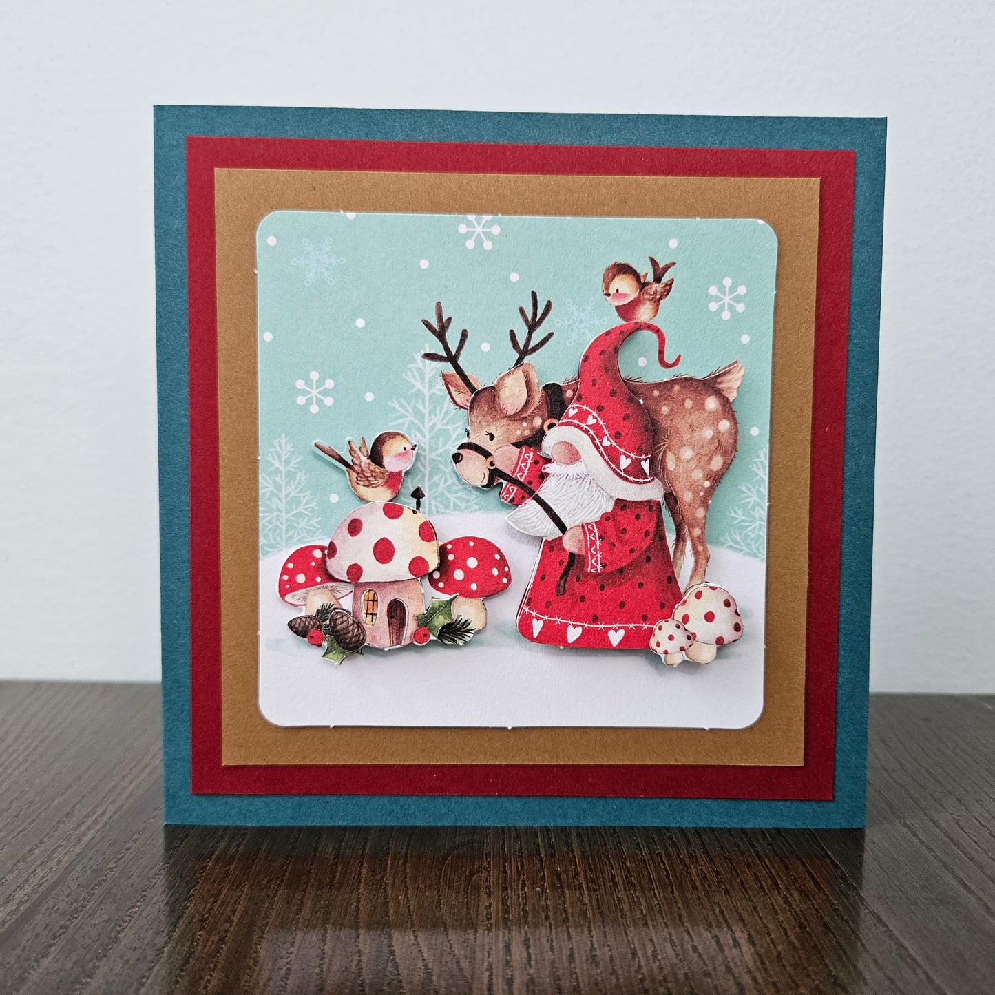 A square handmade greeting card featuring a festive Christmas illustration of a deer, a monkey, and a small house on a snowy ground, layered over red, kraft brown, and teal cardstock borders on a dark wooden surface.