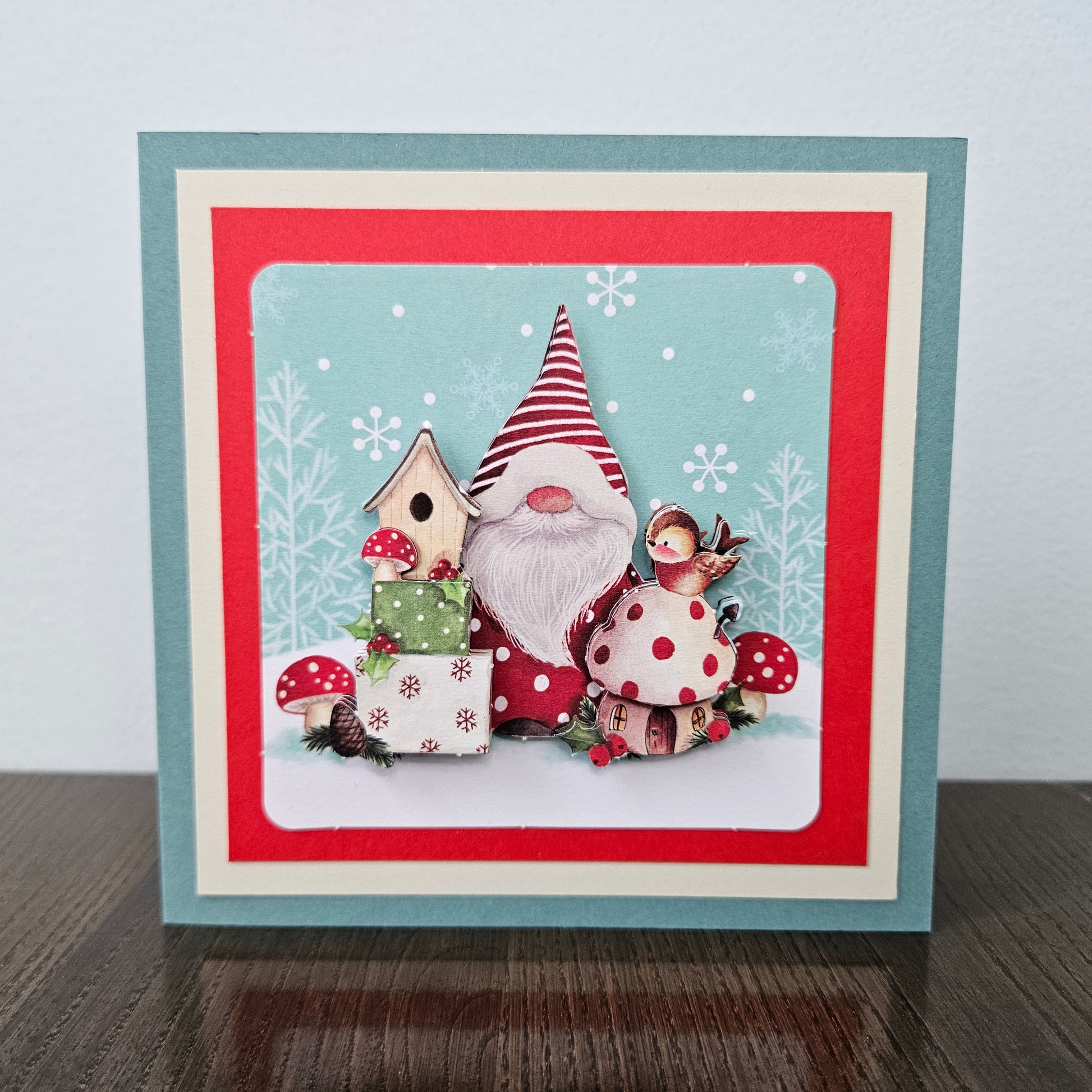A square handmade greeting card featuring a whimsical gnome scene with red-and-white polka dots, a birdhouse, gift boxes, mushrooms, and a small bird, set against a light blue snowy background. The card has layered borders in teal, red, and cream, and is photographed standing on a dark wooden surface against a light gray wall.
