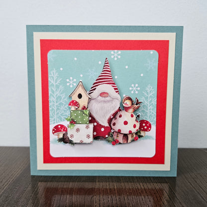 A square handmade greeting card featuring a whimsical gnome scene with red-and-white polka dots, a birdhouse, gift boxes, mushrooms, and a small bird, set against a light blue snowy background. The card has layered borders in teal, red, and cream, and is photographed standing on a dark wooden surface against a light gray wall.