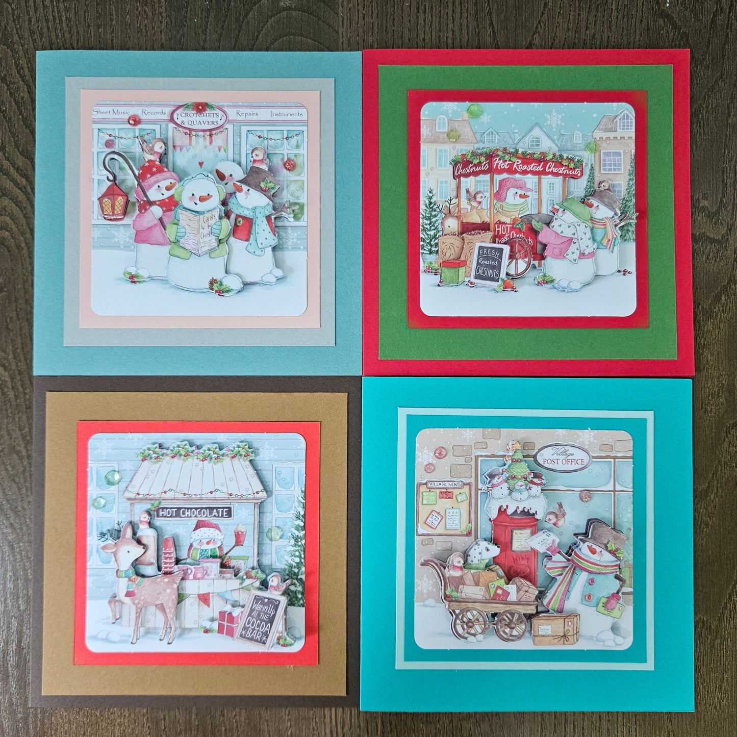 Four handmade square Christmas cards arranged in a 2x2 grid on a dark wood background. Each card features a different holiday illustration with layered colored borders: teal, red, gold, and teal frames around festive scenes with snowmen, Santa, a hot chocolate stand, and a gift shop.