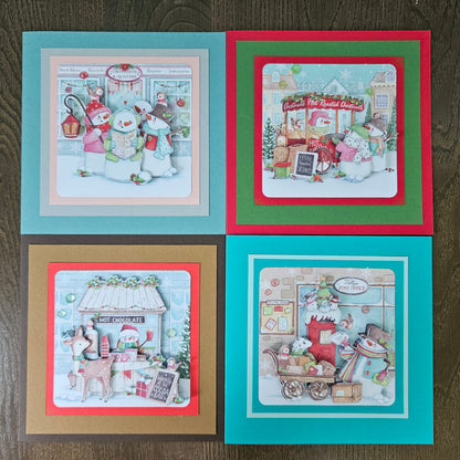 Four handmade square Christmas cards arranged in a 2x2 grid on a dark wood background. Each card features a different holiday illustration with layered colored borders: teal, red, gold, and teal frames around festive scenes with snowmen, Santa, a hot chocolate stand, and a gift shop.