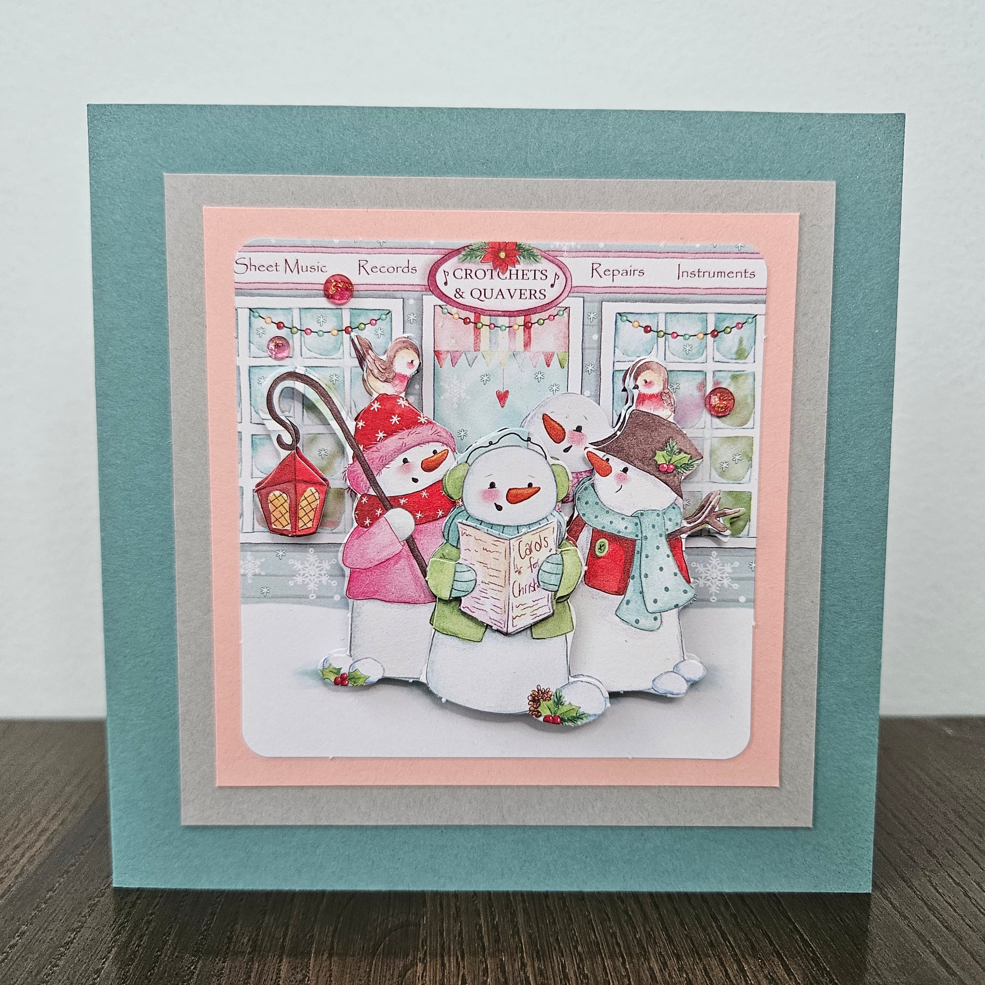 A square handmade greeting card featuring a festive winter illustration of three snowmen reading in front of a music shop. The card has layered borders in teal, light pink, and gray, and is displayed standing on a dark wooden surface against a light gray wall.