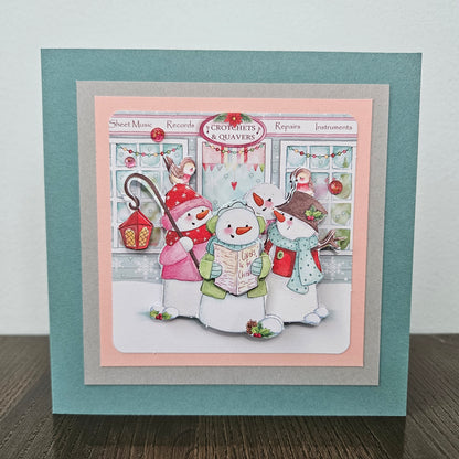 A square handmade greeting card featuring a festive winter illustration of three snowmen reading in front of a music shop. The card has layered borders in teal, light pink, and gray, and is displayed standing on a dark wooden surface against a light gray wall.
