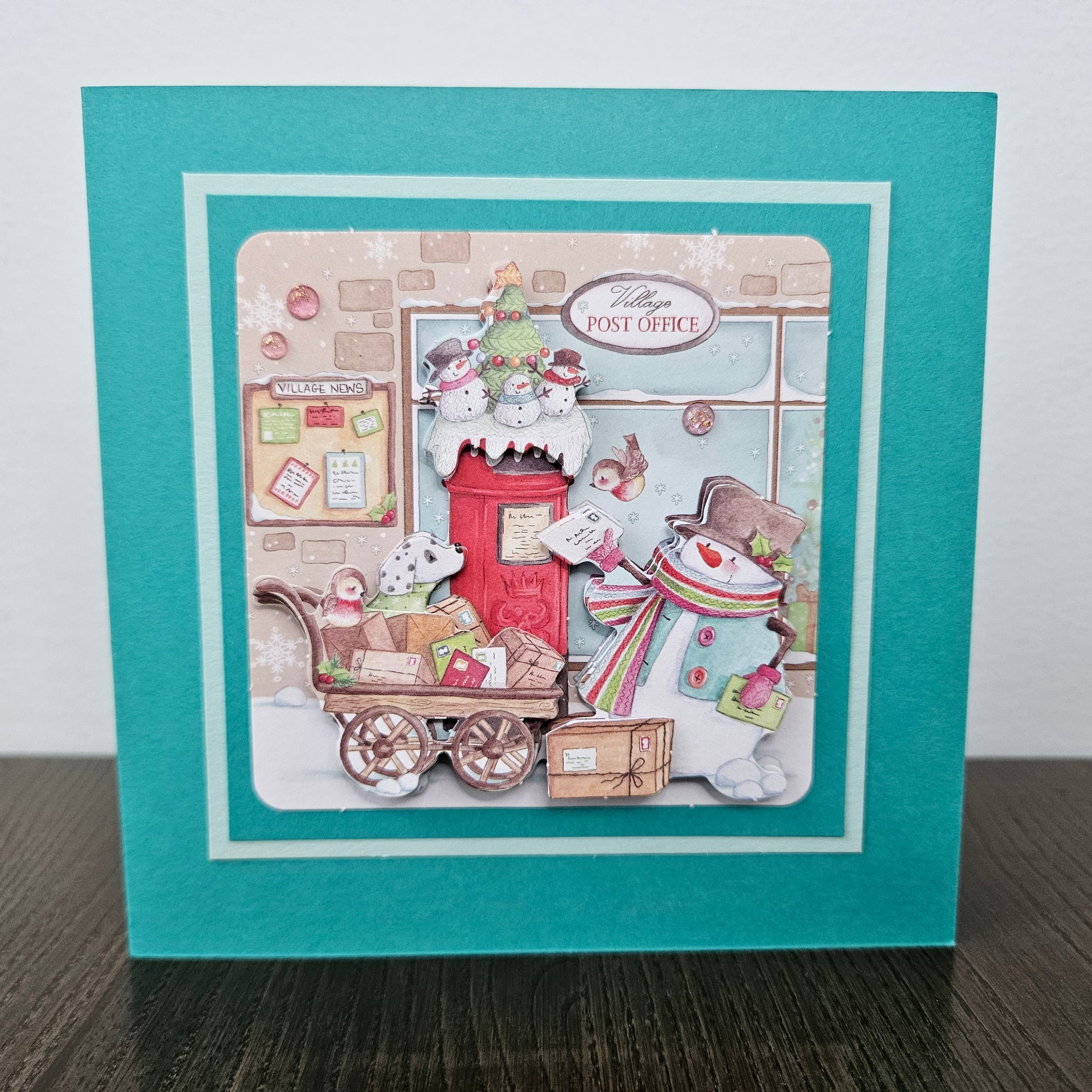 A handmade layered card featuring a winter post office scene. The card has a teal outer border, a white inner border, and a central die-cut illustration showing a red post box, a snowman in a striped scarf, a snowball cart, and a Christmas tree on a snowy street.