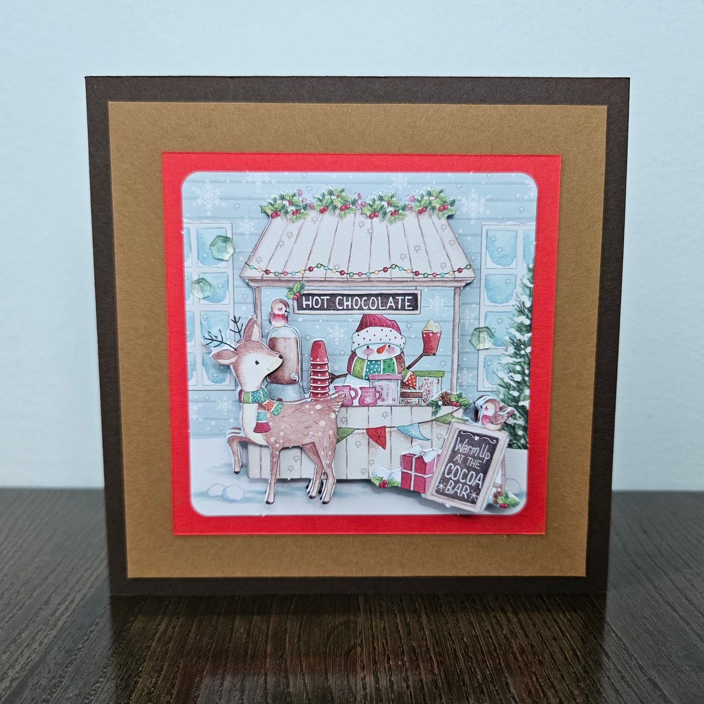 A square handmade greeting card featuring a festive Christmas hot chocolate bar scene, displayed on a dark wooden surface against a light blue wall. The card has a dark brown outer border, a wide tan mat, and a red inner border framing a detailed illustration of a decorated hot chocolate shop with a reindeer and a snowman.