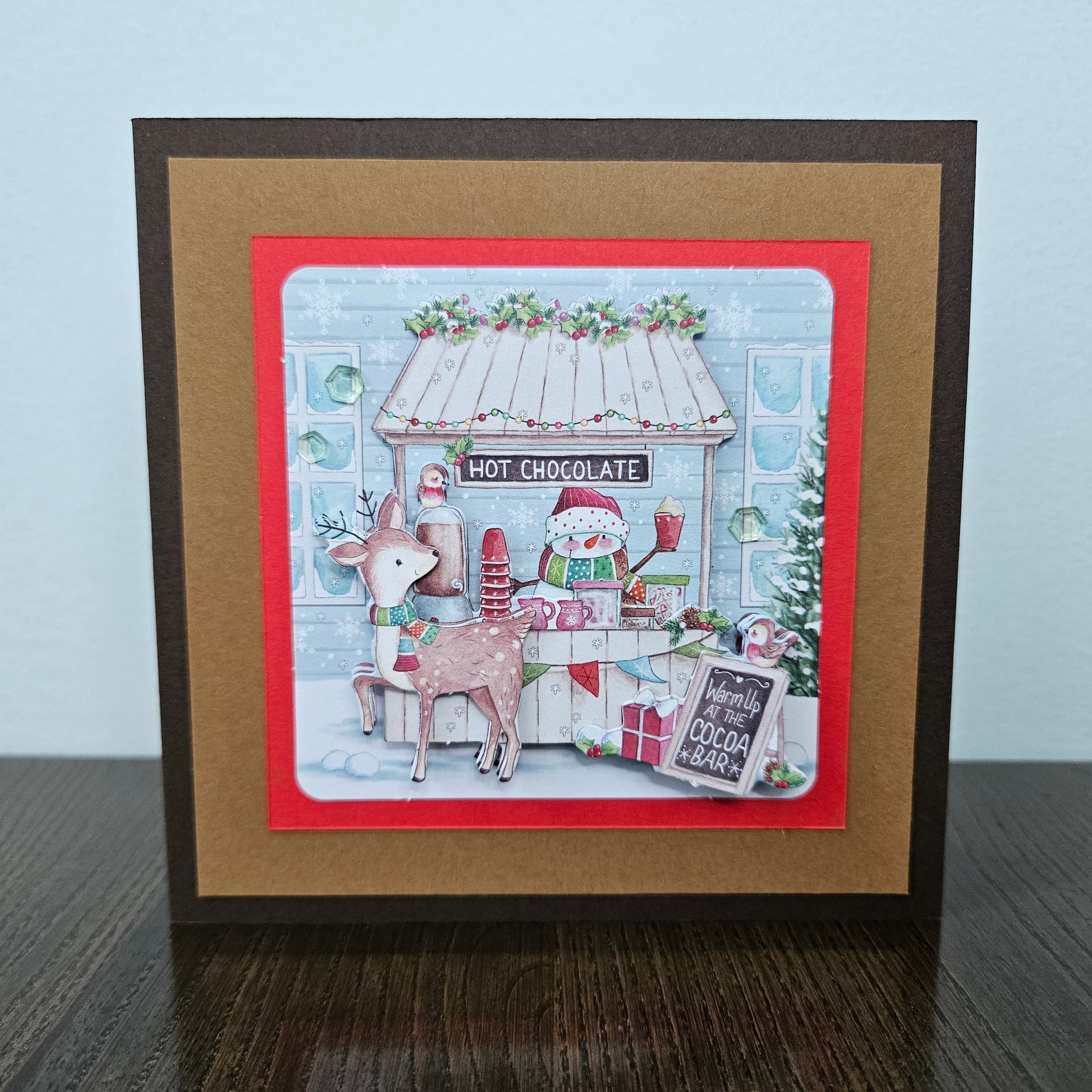 A square handmade greeting card featuring a festive Christmas hot chocolate bar scene, displayed on a dark wooden surface against a light blue wall. The card has a dark brown outer border, a wide tan mat, and a red inner border framing a detailed illustration of a decorated hot chocolate shop with a reindeer and a snowman.