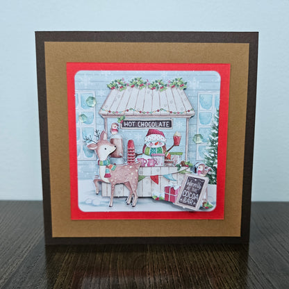 A square handmade greeting card featuring a festive Christmas hot chocolate bar scene, displayed on a dark wooden surface against a light blue wall. The card has a dark brown outer border, a wide tan mat, and a red inner border framing a detailed illustration of a decorated hot chocolate shop with a reindeer and a snowman.