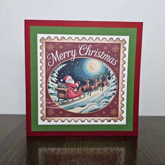 A square Christmas greeting card standing upright on a dark wooden surface against a light gray wall. The card has a red border, a green inner border, and a central illustrated scene showing Santa Claus in a sleigh pulled by reindeer under a full moon, with the words “Merry Christmas” across the top.