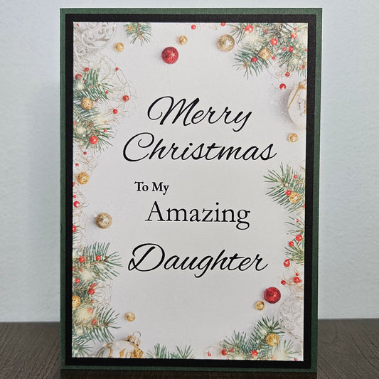 A rectangular Christmas greeting card standing upright on a dark wooden surface against a light gray wall. The card has a dark green border and reads 'Merry Christmas To My Amazing Daughter' in black script and serif text, surrounded by holiday pine branches, red berries, and gold ornaments.