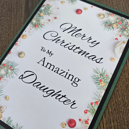 A close-up of a Christmas greeting card with the text 'Merry Christmas To My Amazing Daughter' in black script, decorated with green pine branches, red berries, and gold ornaments on a white background, placed on a wooden surface.