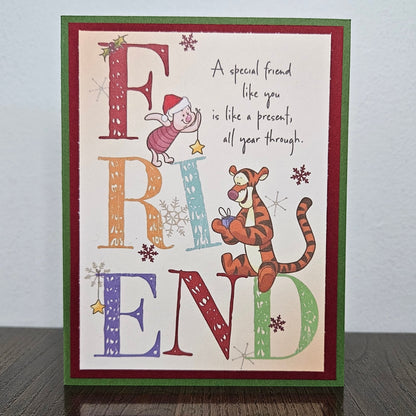 A handmade Christmas greeting card featuring the word 'FRIEND' in large decorative letters with Piglet and Tigger illustrations, standing on a dark wooden surface against a light gray wall.