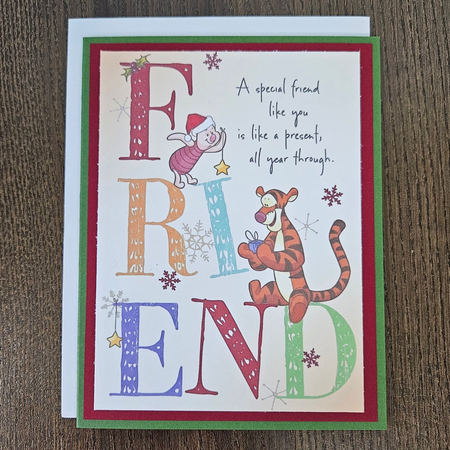 A square greeting card featuring a Christmas-themed 'Friend' design, placed on a dark wood surface. The card has a white center with colorful decorative letters spelling FRIEND, illustrated characters in holiday attire, snowflakes, and a quote.