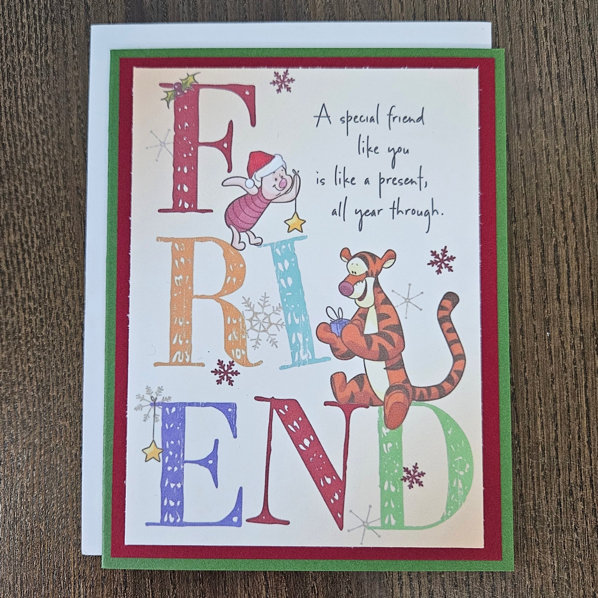 A square greeting card featuring a Christmas-themed 'Friend' design, placed on a dark wood surface. The card has a white center with colorful decorative letters spelling FRIEND, illustrated characters in holiday attire, snowflakes, and a quote.