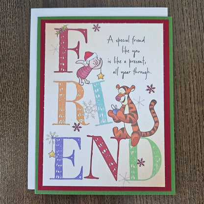 A square greeting card featuring a Christmas-themed 'Friend' design, placed on a dark wood surface. The card has a white center with colorful decorative letters spelling FRIEND, illustrated characters in holiday attire, snowflakes, and a quote.
