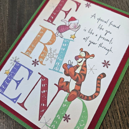 A square Christmas greeting card featuring the word 'FRIEND' in large decorative letters, with Winnie the Pooh and Tigger illustrations and a handwritten message on a white background, photographed on a wooden surface.