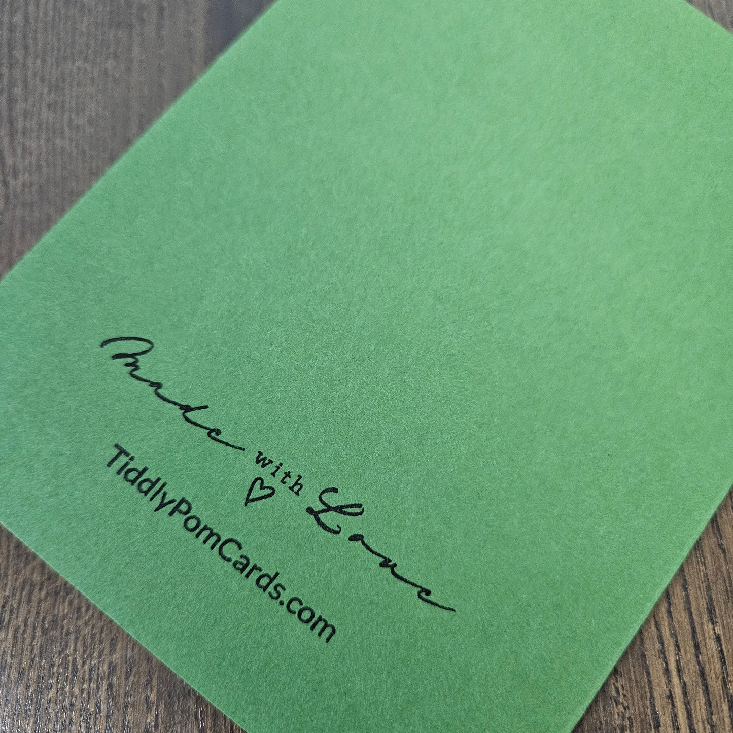A green textured card with black printed text reading 'Made with Love' and 'TiddyPomCards.com' resting on a dark wood surface.