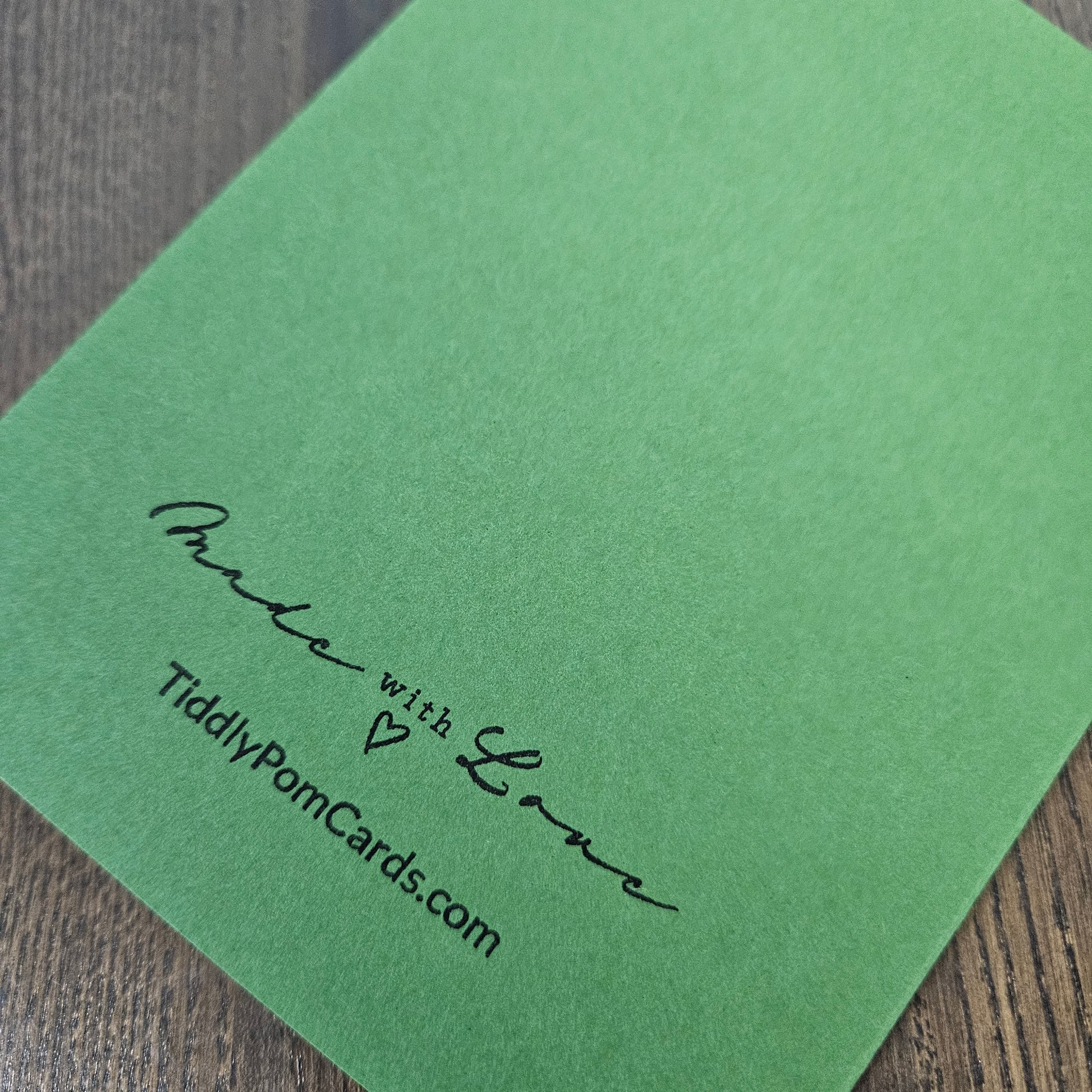 A green textured card with black printed text reading 'Made with Love' and 'TiddyPomCards.com' resting on a dark wood surface.