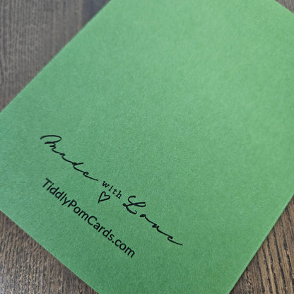 A green textured card with black printed text reading 'Made with Love' and 'TiddyPomCards.com' resting on a dark wood surface.