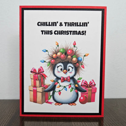 A handmade Christmas greeting card featuring a cute cartoon penguin wearing a messy hairdo of Christmas lights, standing on a dark wooden table against a light gray wall. The card has a red border, black inner border, and the text 'CHILLIN' & THRILLIN' THIS CHRISTMAS!' at the top.