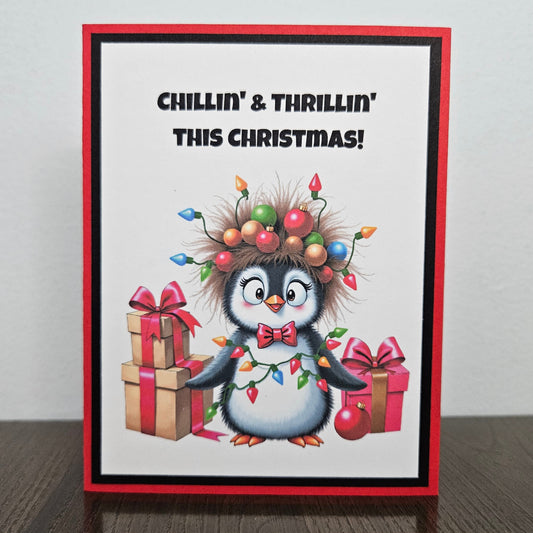 A handmade Christmas greeting card featuring a cute cartoon penguin wearing a messy hairdo of Christmas lights, standing on a dark wooden table against a light gray wall. The card has a red border, black inner border, and the text 'CHILLIN' & THRILLIN' THIS CHRISTMAS!' at the top.
