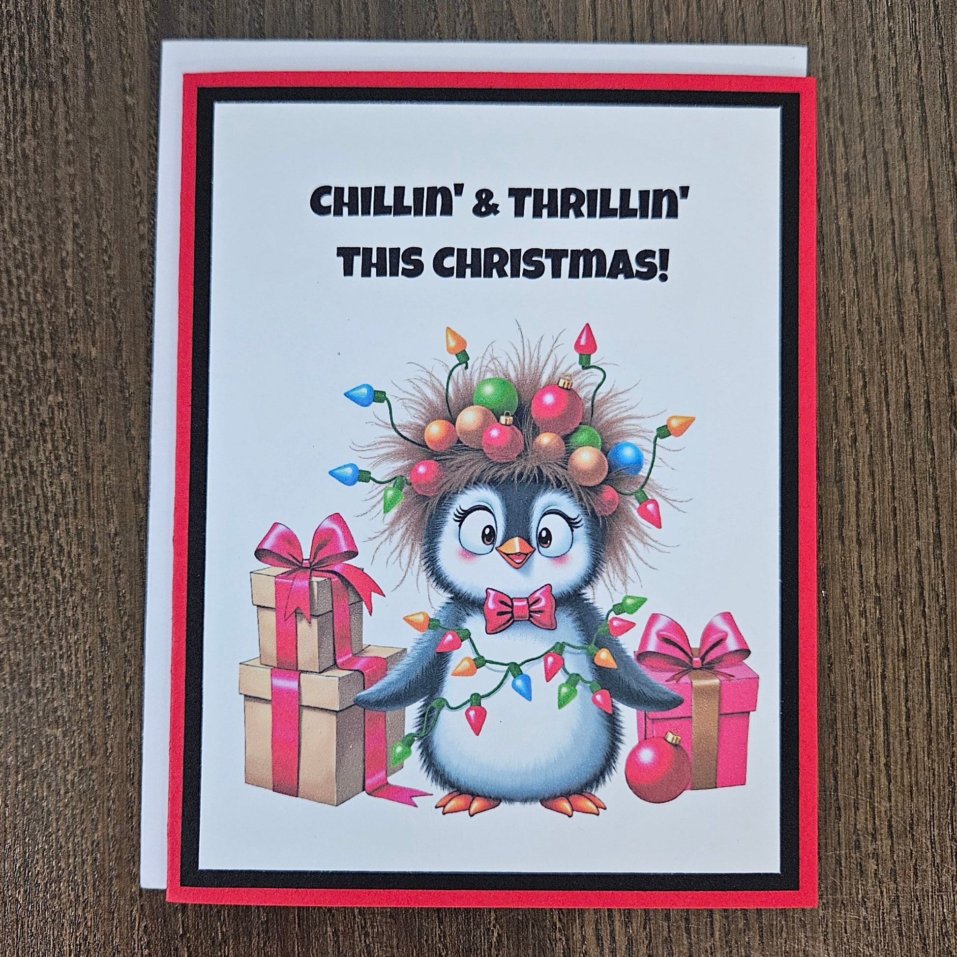 A Christmas greeting card featuring a cartoon penguin wearing colorful holiday lights and ornaments, standing beside wrapped gifts. The card reads 'CHILLIN' & THRILLIN' THIS CHRISTMAS!' and is photographed on a brown wood surface.