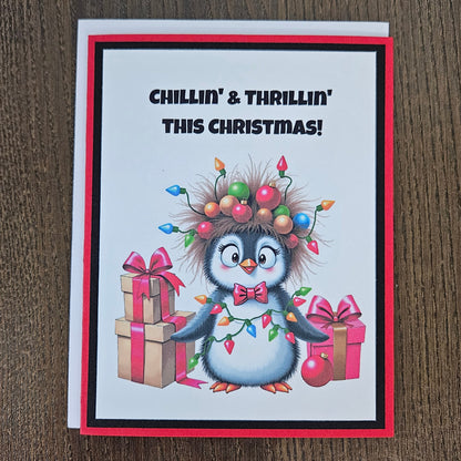 A Christmas greeting card featuring a cartoon penguin wearing colorful holiday lights and ornaments, standing beside wrapped gifts. The card reads 'CHILLIN' & THRILLIN' THIS CHRISTMAS!' and is photographed on a brown wood surface.