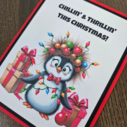 A square Christmas greeting card featuring a cartoon penguin surrounded by presents, holiday lights, and the text 'CHILLIN' & THRILLIN' THIS CHRISTMAS!' on a white background with a red border, photographed on a wooden surface.