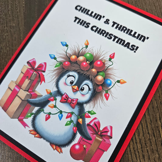A square Christmas greeting card featuring a cartoon penguin surrounded by presents, holiday lights, and the text 'CHILLIN' & THRILLIN' THIS CHRISTMAS!' on a white background with a red border, photographed on a wooden surface.