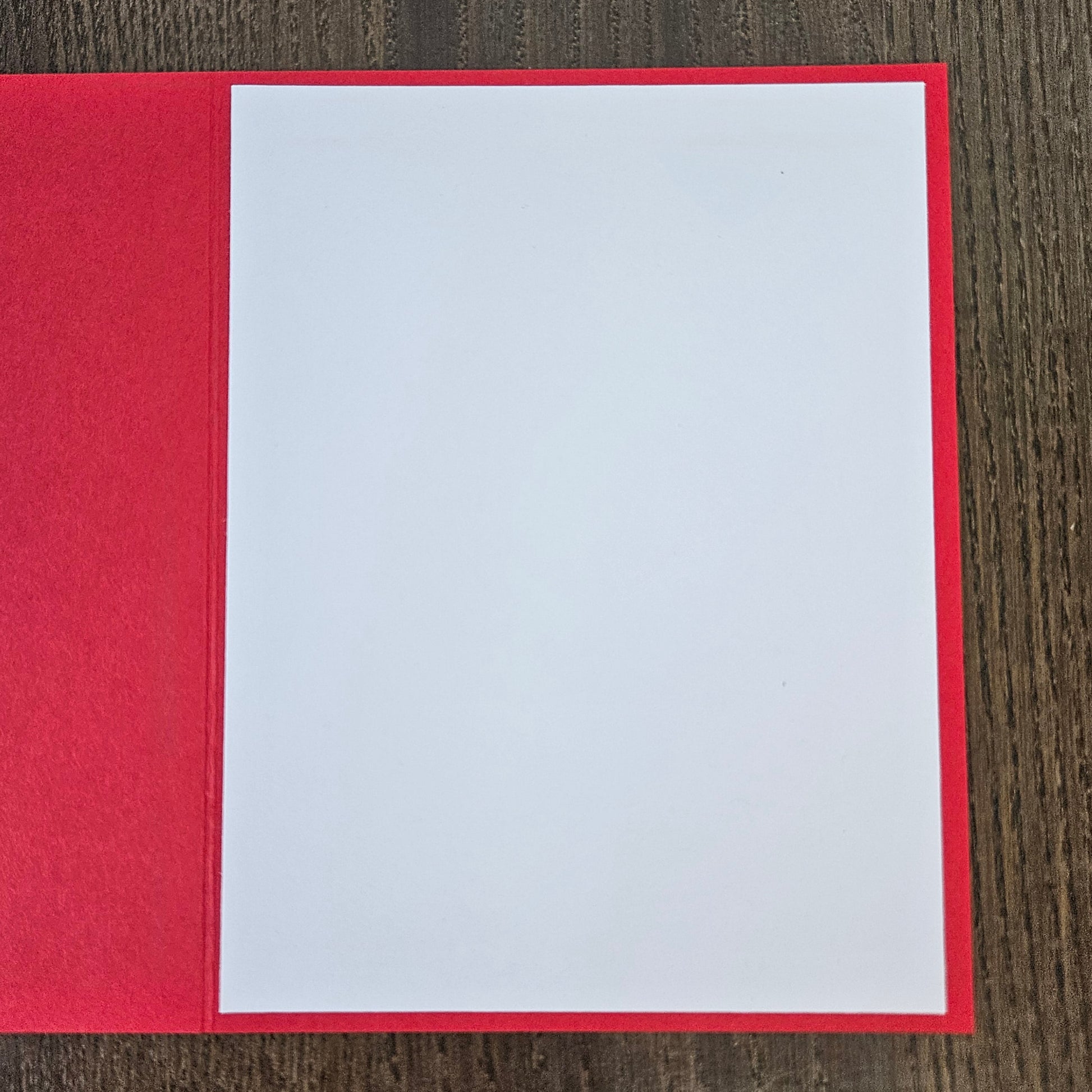 A red greeting card or notebook cover with a blank white insert page, photographed from above on a dark wood surface.
