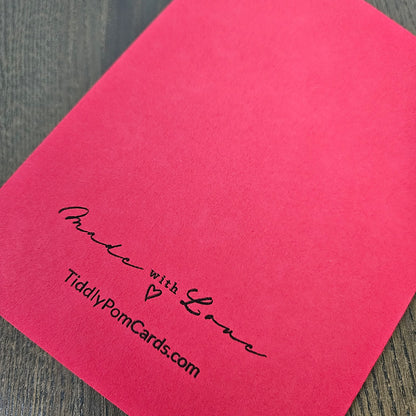 A bright pink square card with black printed text reading 'Make with Love' and 'TiddlyPomCards.com' placed on a dark wood surface.
