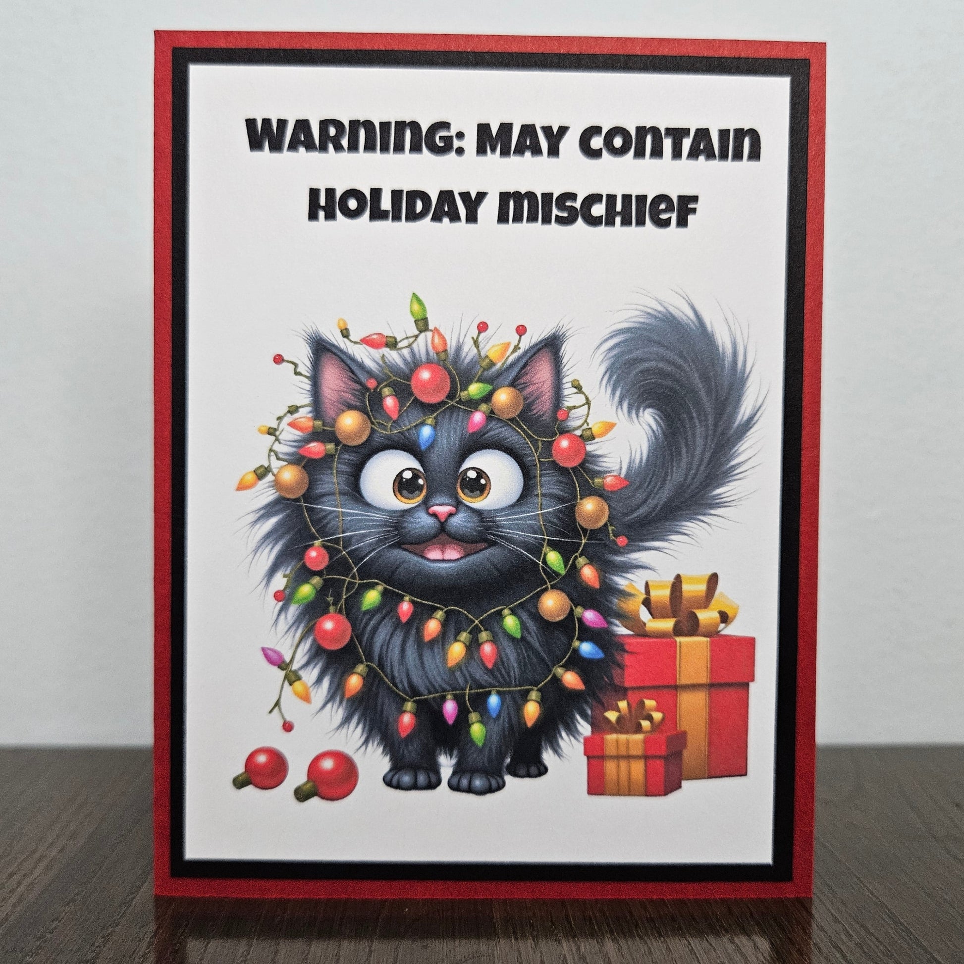 Front of a funny handmade A2 Christmas card featuring a black cat tangled in Christmas lights with text reading Warning May Contain Holiday Mischief framed in red and green.