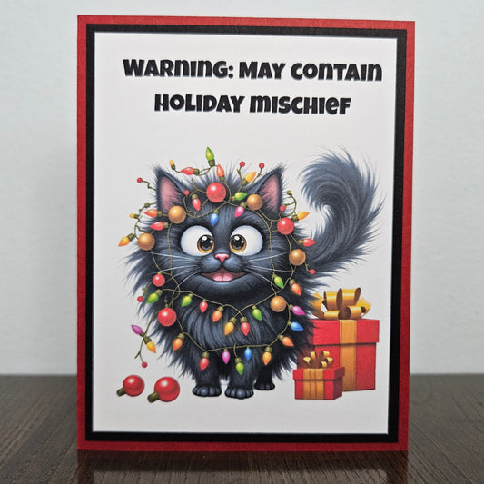 Front of a funny handmade A2 Christmas card featuring a black cat tangled in Christmas lights with text reading Warning May Contain Holiday Mischief framed in red and green.