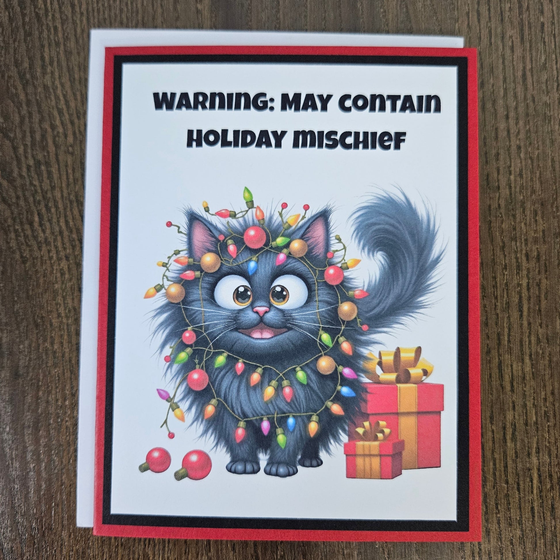 A greeting card featuring a cartoon fluffy gray cat wrapped in multicolored Christmas lights, with the text 'WARNING: MAY CONTAIN HOLIDAY MISCHIEF' at the top. The card has a red border and is photographed on a brown wood surface.