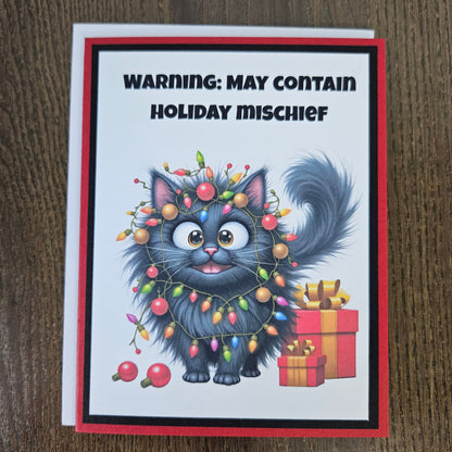 A greeting card featuring a cartoon fluffy gray cat wrapped in multicolored Christmas lights, with the text 'WARNING: MAY CONTAIN HOLIDAY MISCHIEF' at the top. The card has a red border and is photographed on a brown wood surface.