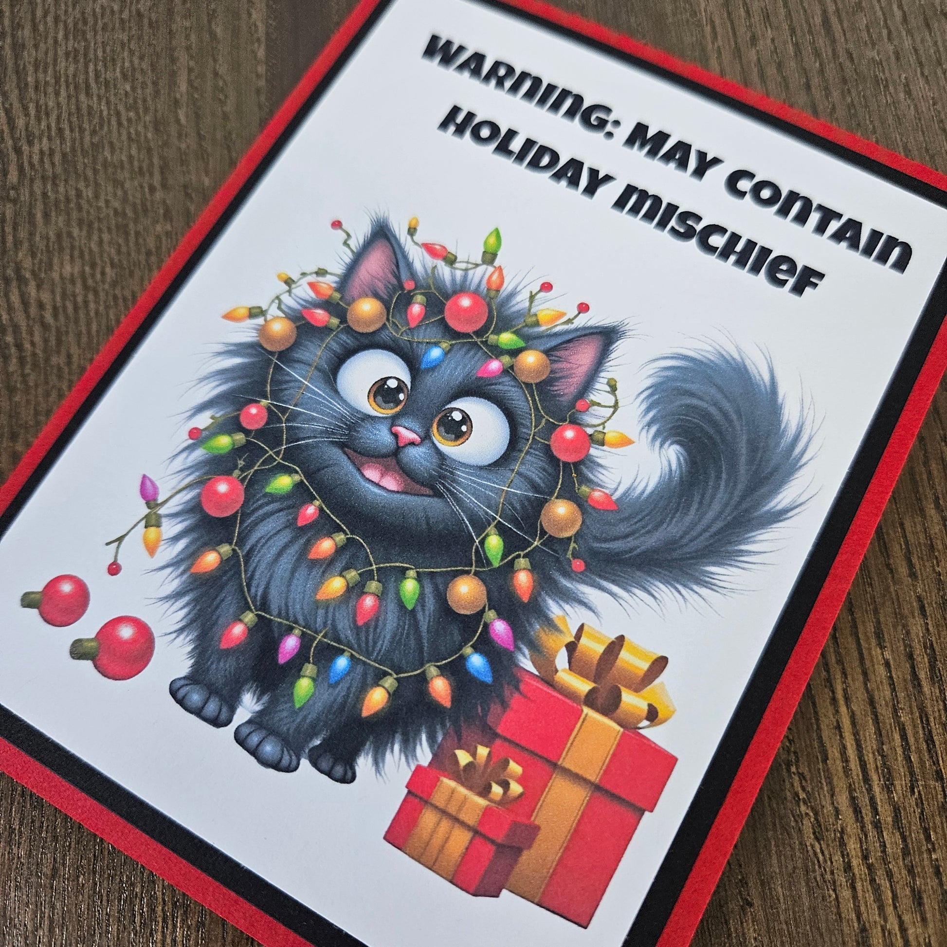 A square greeting card featuring a cartoon fluffy black cat wrapped in multicolored Christmas lights, with the text 'WARNING: MAY CONTAIN HOLIDAY MISCHIEF' at the top. The card has a red border and is photographed on a wooden surface.