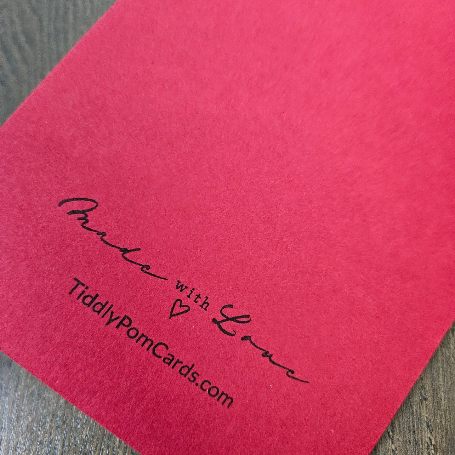 A close-up of a bright pink textured card with black printed text reading 'Made with Love' and 'TiddlyPomCards.com' on a gray wood surface.
