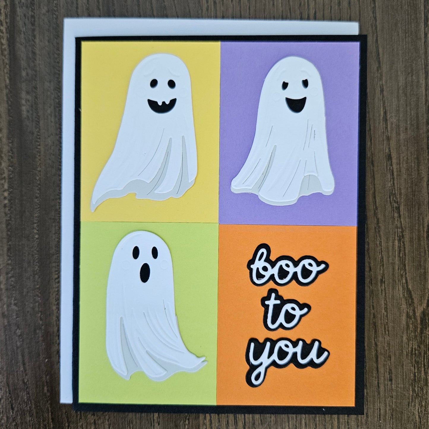 A square Halloween greeting card featuring a four-panel ghost design in yellow, purple, green, and orange, with the text 'boo to you' on the bottom right, photographed on a dark wood surface.