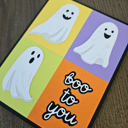 A square Halloween greeting card with a four-quadrant design in yellow, purple, light green, and orange, featuring three white ghost cutouts and the words 'too to you' in black script with white outlines, photographed on a dark wood surface.