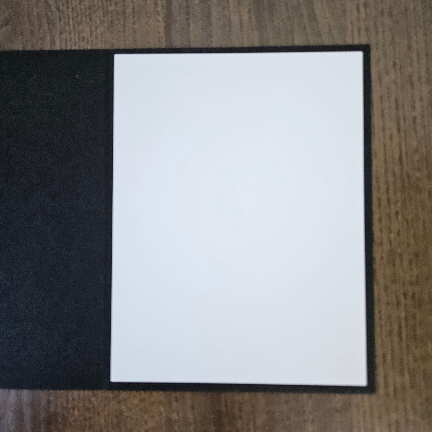 A blank white card or paper insert with a black border, placed on a textured black surface over a wooden table background.
