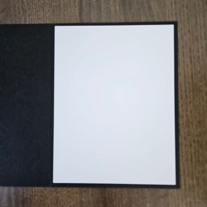 A blank white card or paper insert with a black border, placed on a textured black surface over a wooden table background.