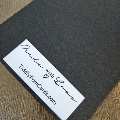 A close-up of a black textured card or envelope with a white rectangular label reading 'Made with Love' and 'TiddlyPornCards.com' on a wooden surface.