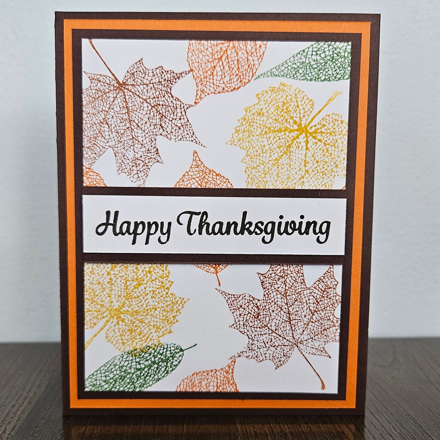 A handmade Thanksgiving greeting card standing upright on a dark wooden surface against a light gray wall. The card features a white background with layered orange and dark brown borders, stamped autumn leaf designs in yellow, orange, and green, and a central black banner reading 'Happy Thanksgiving' in black script.