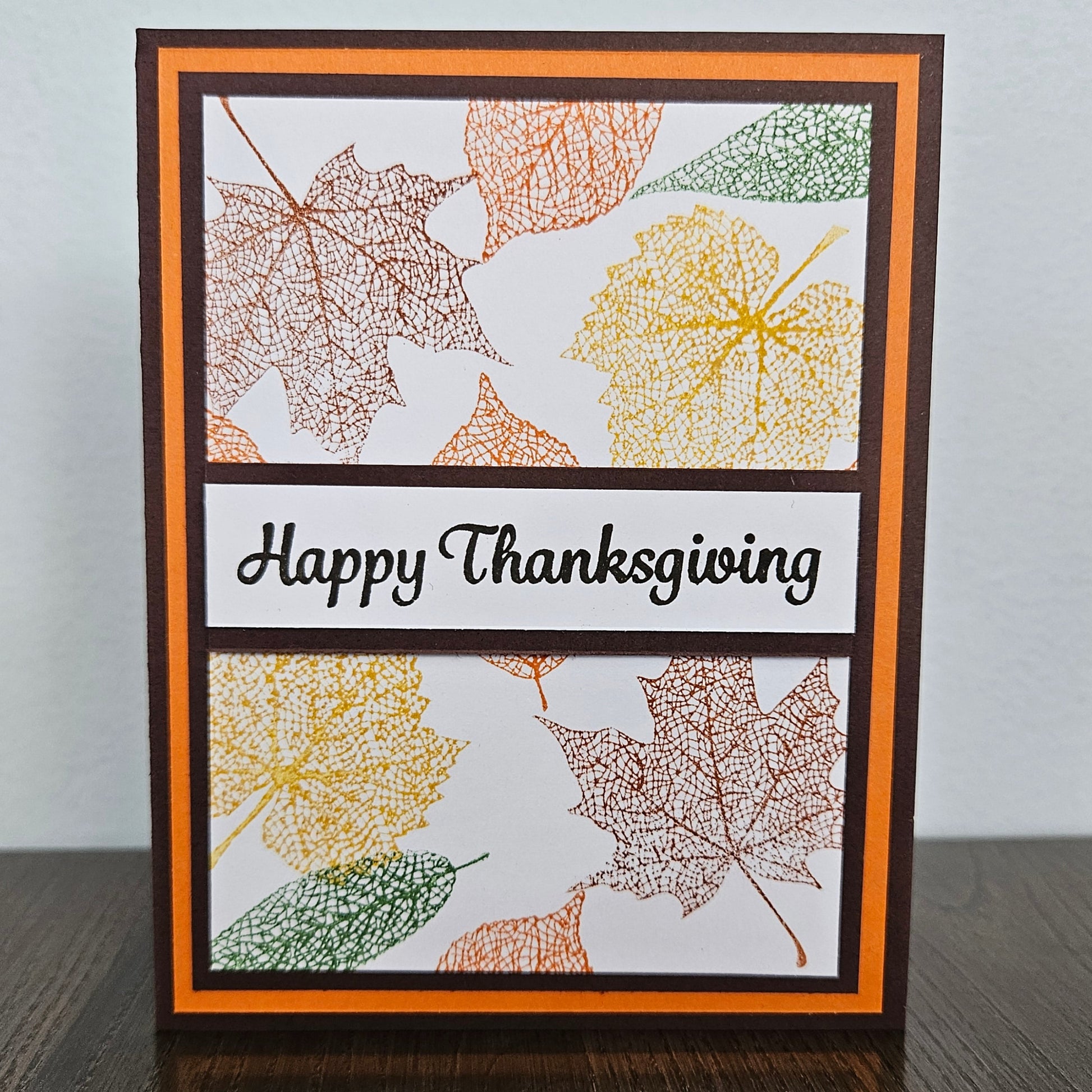 A handmade Thanksgiving greeting card standing upright on a dark wooden surface against a light gray wall. The card features a white background with layered orange and dark brown borders, stamped autumn leaf designs in yellow, orange, and green, and a central black banner reading 'Happy Thanksgiving' in black script.