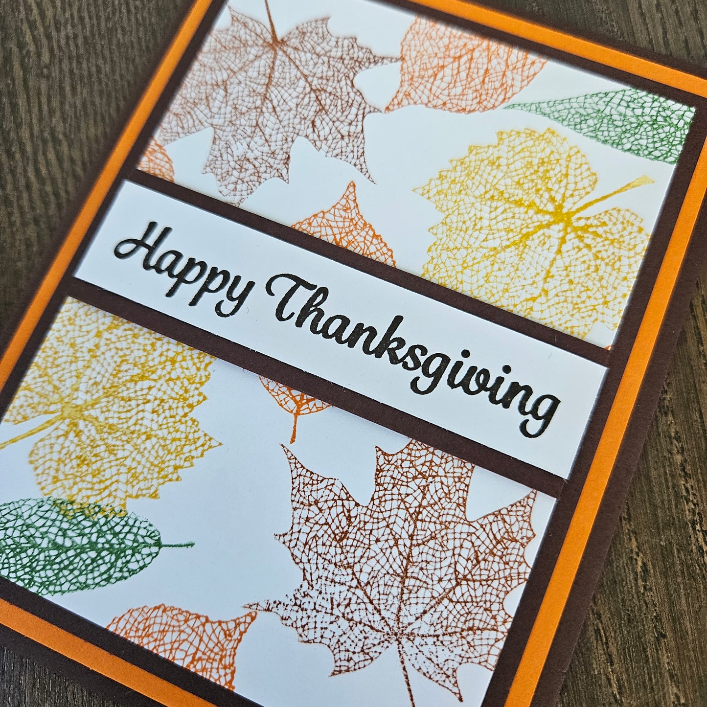 A close-up of a handmade Thanksgiving greeting card featuring a white background with stamped multicolor autumn leaves in yellow, orange, brown, and green. A dark brown banner across the center reads 'Happy Thanksgiving' in black script.
